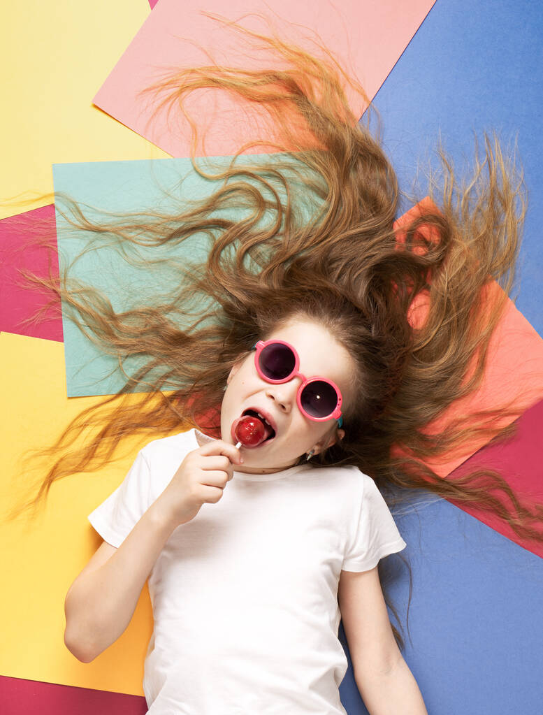 Colores vibrantes: una niña yace sobre un fondo colorido y lame una piruleta
. - Foto, imagen