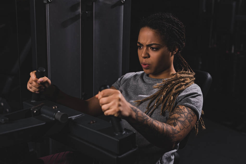 selectieve focus van getatoeëerd Afrikaans Amerikaans meisje met dreadlocks uit te werken op fitness-apparaat - Foto, afbeelding