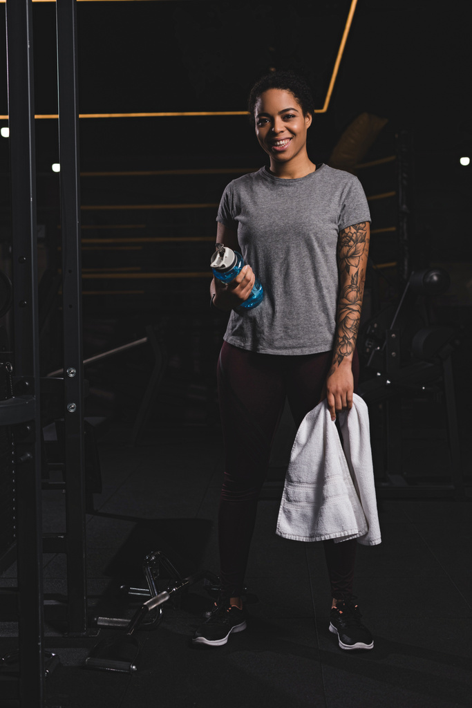 счастливая африканская американка с татуировкой держа спортивную бутылку с водой и стоя в тренажерном зале
  - Фото, изображение