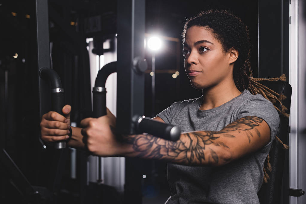 selective focus of young african american girl with tattoo training on gym machine  - Photo, Image