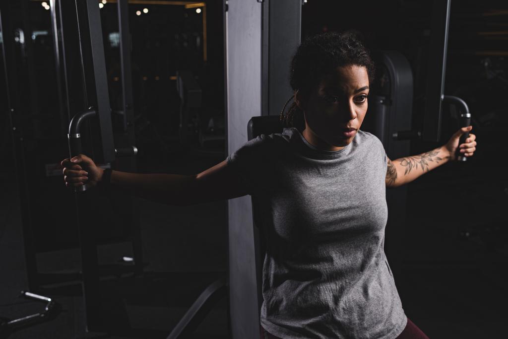  νεαρή Αφροαμερικανή με τατουάζ που εκπαιδεύεται στο μηχάνημα γυμναστικής  - Φωτογραφία, εικόνα