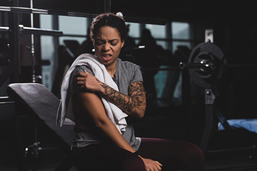 τραυματισμένη Αφροαμερικανή γυναίκα με τατουάζ αγγίζοντας το χέρι, ενώ υποφέρει από πόνο στο γυμναστήριο  - Φωτογραφία, εικόνα