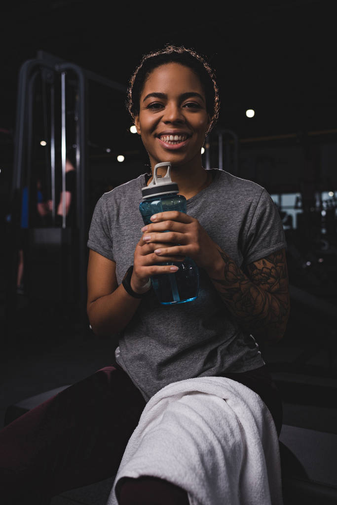 gelukkig en getatoeëerd Afrikaans amerikaanse vrouw met sport fles  - Foto, afbeelding