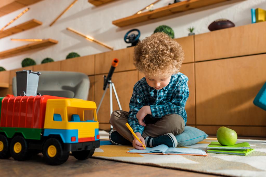 scrittura intelligente per bambini nel taccuino mentre si siede sul pavimento vicino a mela, auto giocattolo e libri
  - Foto, immagini