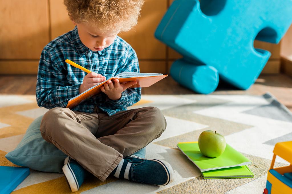 グリーンアップルの近くの床に座っている間にノートに書く賢い子供  - 写真・画像
