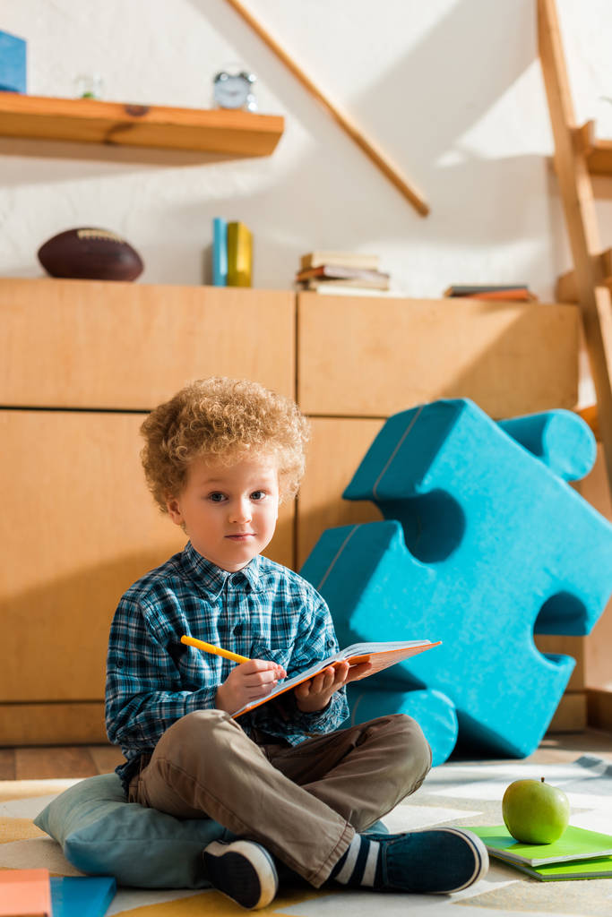intelligentes Kind, das in Notizbuch schreibt, während es auf dem Boden neben Apfel und Büchern sitzt  - Foto, Bild