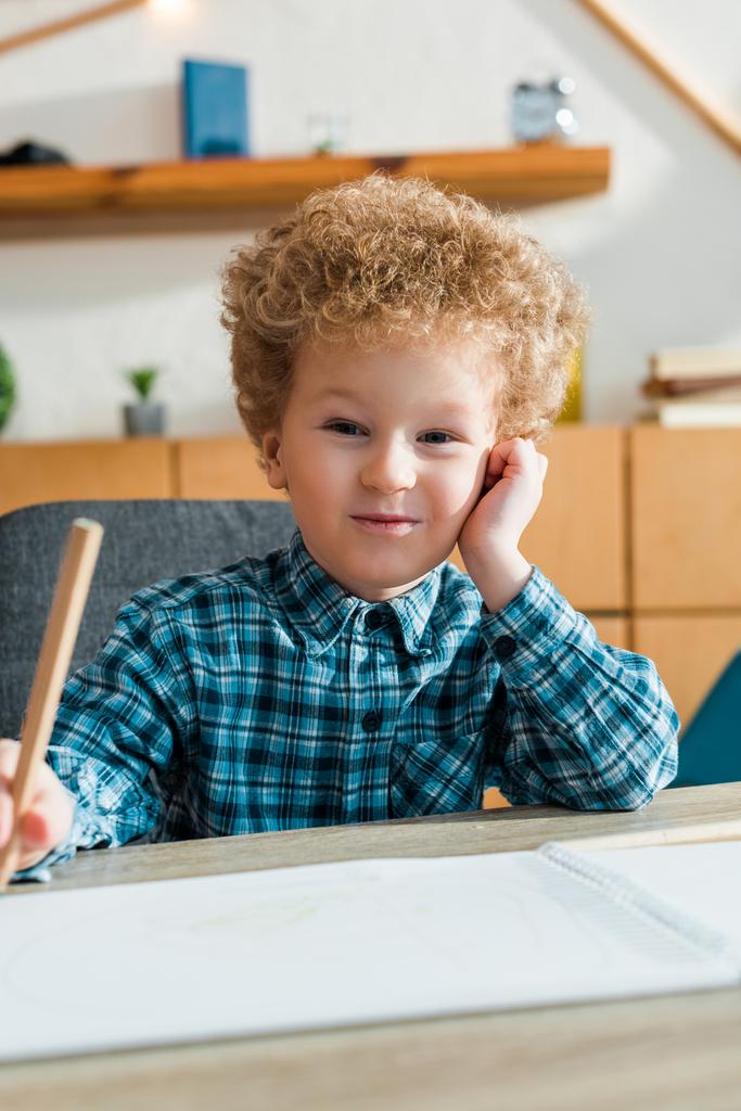 επιλεκτική εστίαση του ευτυχισμένου και σγουρού παιδιού κρατώντας μολύβι κοντά στο κενό χαρτί  - Φωτογραφία, εικόνα