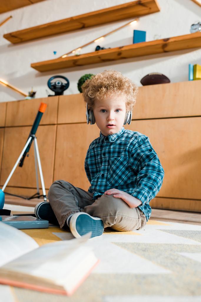 selektywne skupienie zaskoczonego dziecka słuchającego muzyki w słuchawkach bezprzewodowych podczas siedzenia na podłodze  - Zdjęcie, obraz