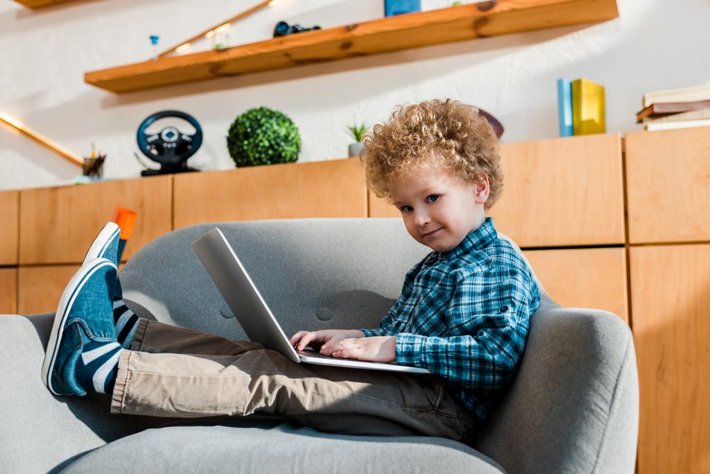 Mutlu çocuk koltukta oturuyor ve dizüstü bilgisayar kullanıyor.  - Fotoğraf, Görsel
