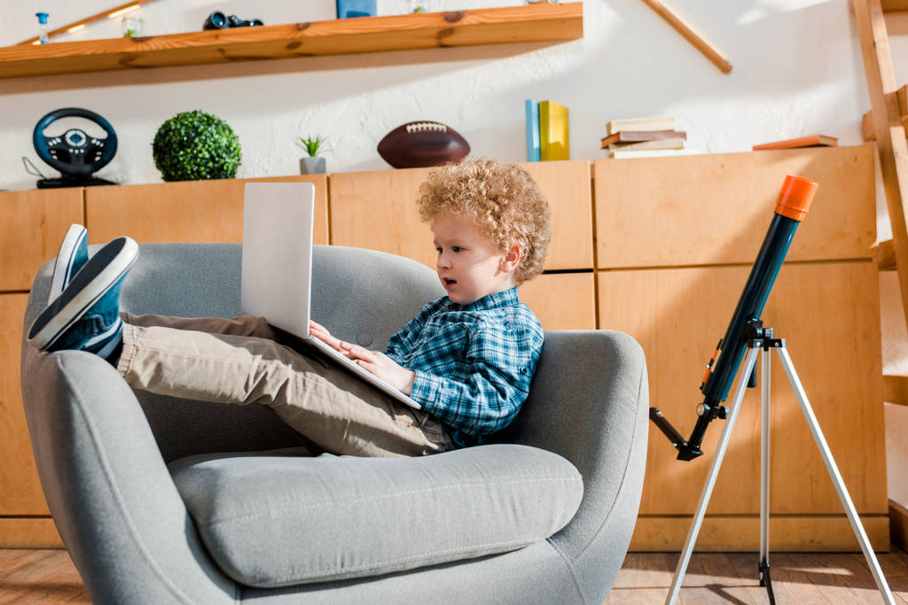 Kıvırcık çocuk koltukta oturuyor ve dizüstü bilgisayar kullanıyor.  - Fotoğraf, Görsel