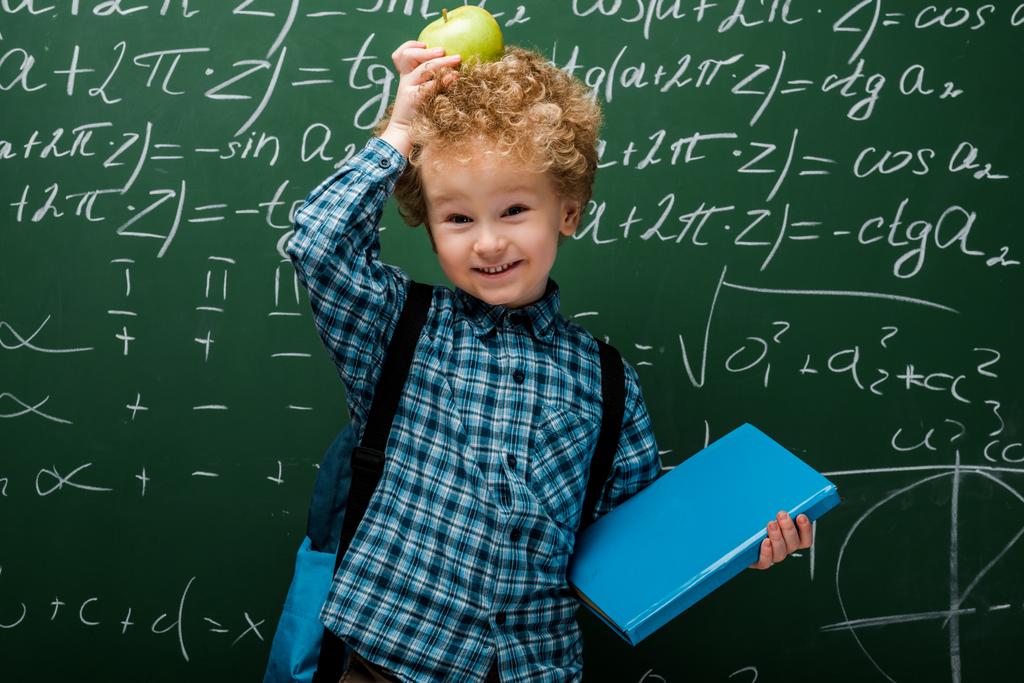 szczęśliwy dzieciak trzyma jabłko i książki w pobliżu tablicy z wzorów matematycznych  - Zdjęcie, obraz