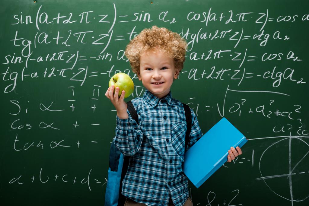 χαρούμενο παιδί κρατώντας το μήλο και το βιβλίο κοντά στο chalkboard με μαθηματικούς τύπους  - Φωτογραφία, εικόνα
