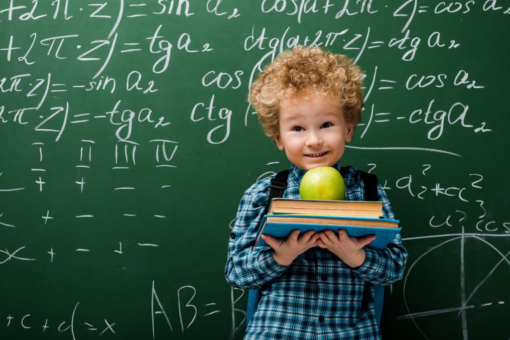 Matematiksel formüllerle dolu karatahtanın yanında kitap ve elma tutan neşeli çocuk.  - Fotoğraf, Görsel