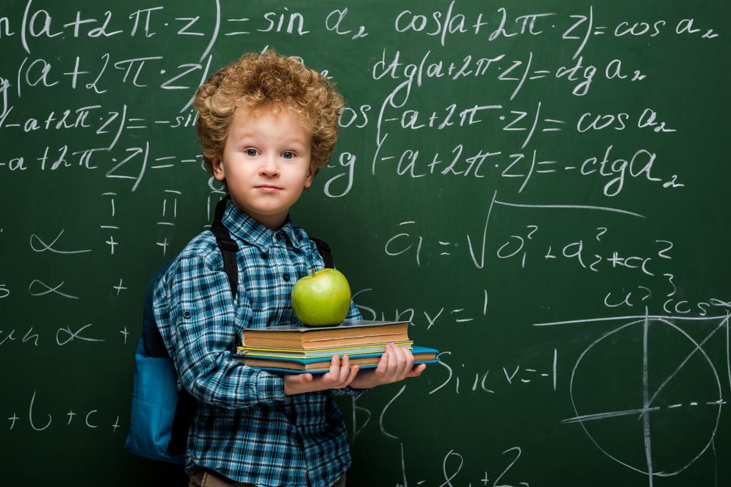 niño rizado sosteniendo libros y manzana fresca cerca de pizarra con fórmulas matemáticas
  - Foto, imagen