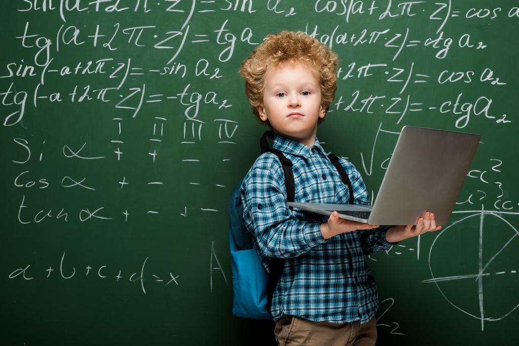 σγουρό παιδί κρατώντας φορητό υπολογιστή κοντά στο chalkboard με μαθηματικούς τύπους  - Φωτογραφία, εικόνα