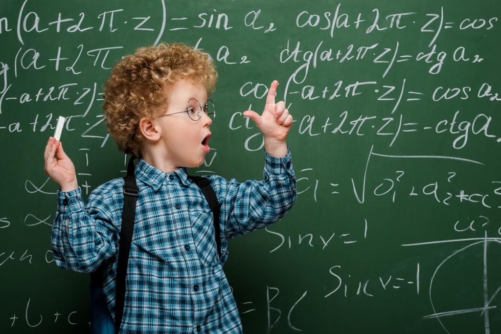 niño sorprendido en gafas haciendo gestos mientras sostiene tiza cerca de pizarra con fórmulas matemáticas
  - Foto, Imagen