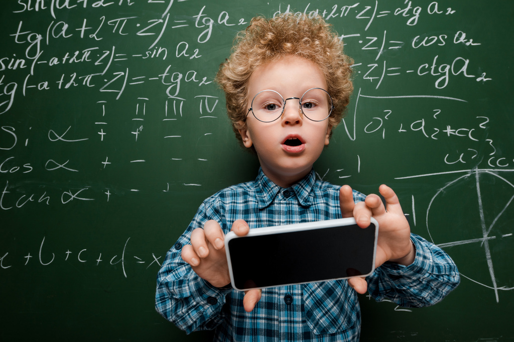 chytré dítě v brýlích drží smartphone s prázdnou obrazovkou v blízkosti tabuli s matematickými vzorci  - Fotografie, Obrázek