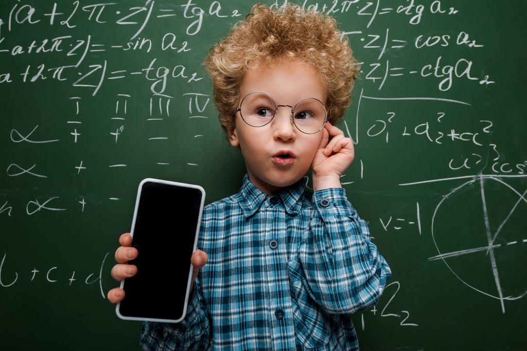 smart child holding smartphone with blank screen and touching glasses near chalkboard with mathematical formulas  - Photo, Image