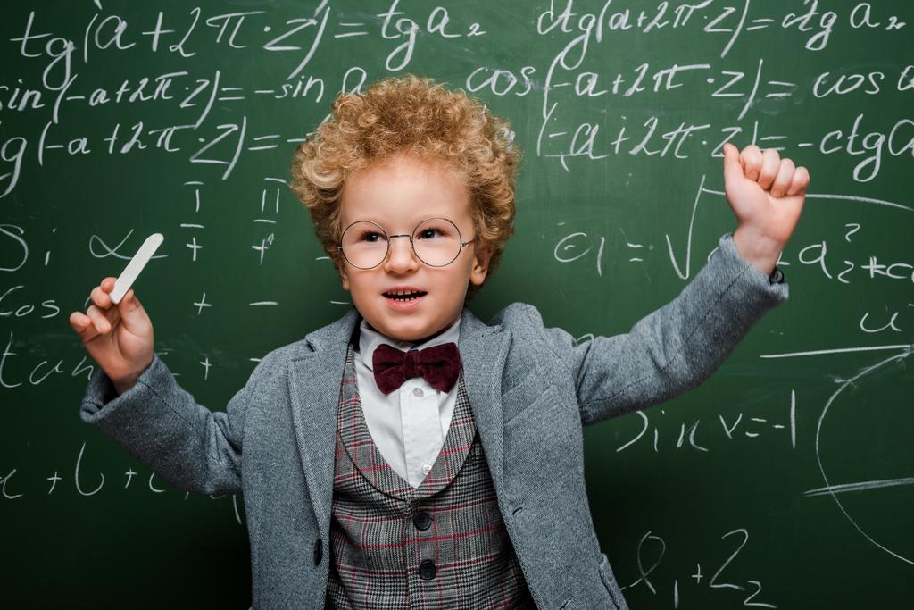 niño inteligente en traje con pajarita sosteniendo tiza cerca de pizarra con fórmulas matemáticas
  - Foto, Imagen