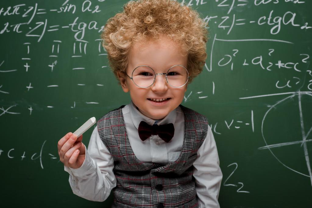 έξυπνο παιδί με κοστούμι και παπιγιόν κρατώντας κιμωλία κοντά σε μαυροπίνακα με μαθηματικούς τύπους  - Φωτογραφία, εικόνα