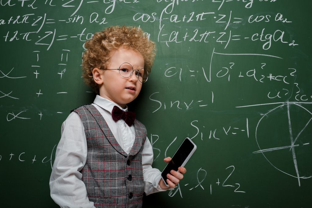 niño disgustado con traje y pajarita sosteniendo teléfono inteligente con pantalla en blanco cerca de pizarra con fórmulas matemáticas
  - Foto, Imagen