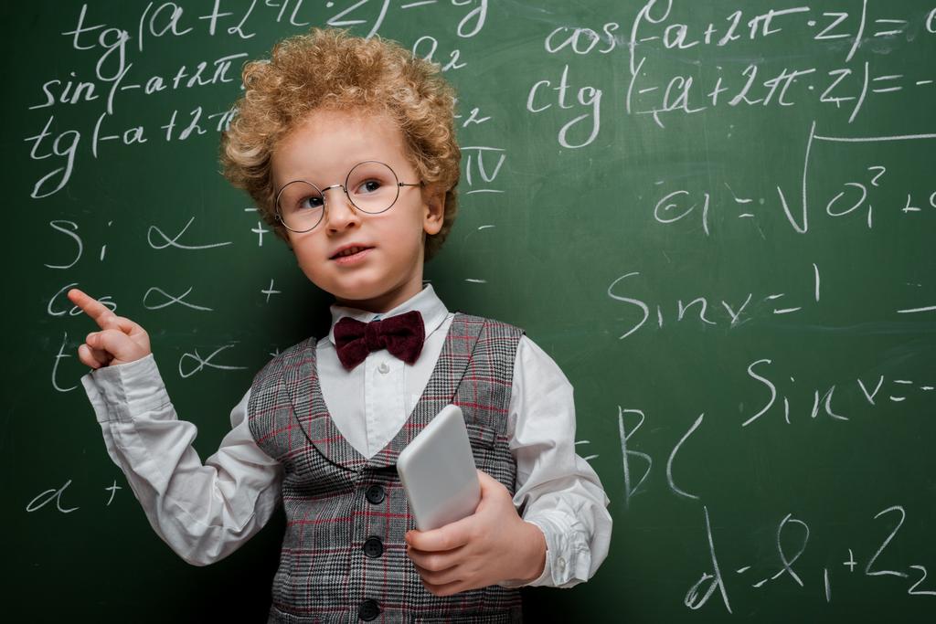 smart child in suit and bow tie holding smartphone and pointing with finger near chalkboard with mathematical formulas  - Photo, Image