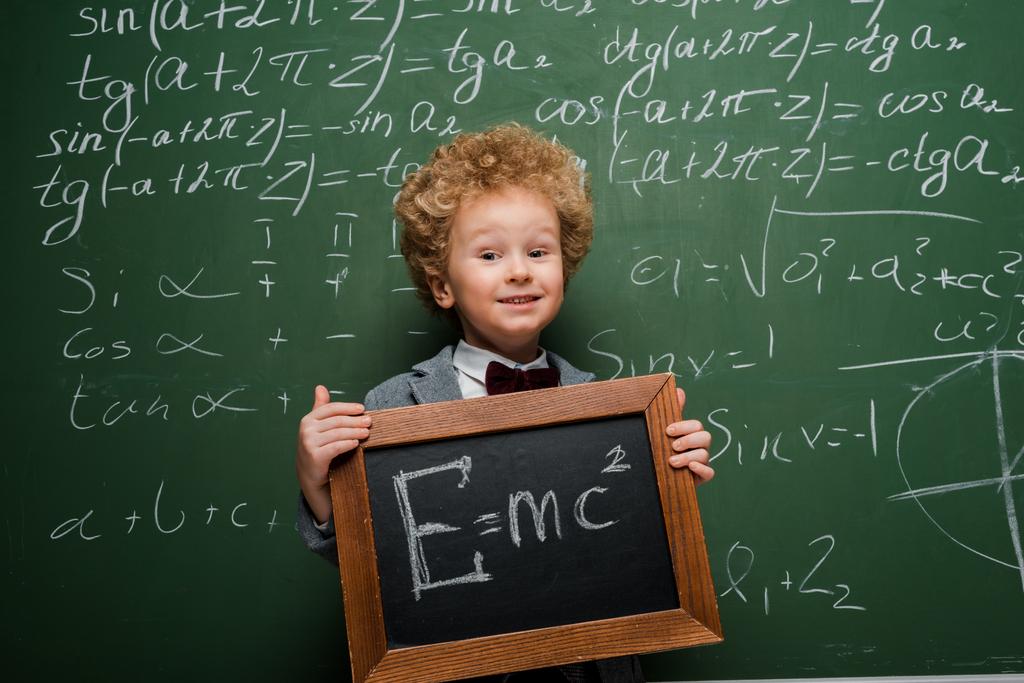 niño feliz en traje y pajarita sosteniendo pequeña pizarra con fórmula cerca de pizarra
  - Foto, Imagen