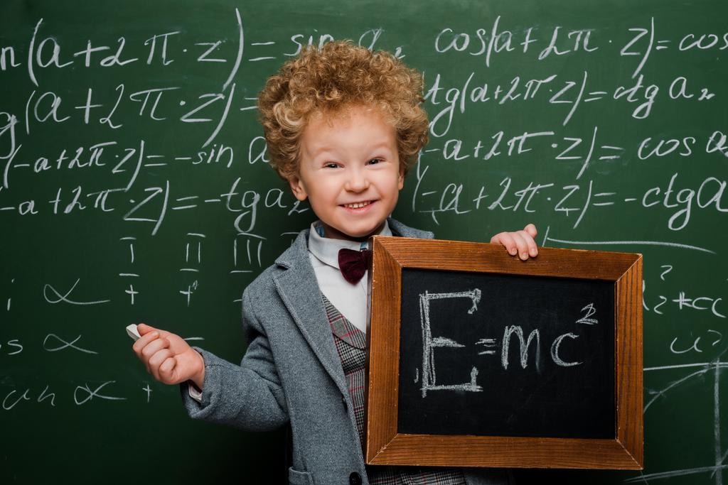 Lächelndes und intelligentes Kind in Anzug und Fliege, das eine kleine Tafel mit Formel in der Nähe der Tafel hält  - Foto, Bild
