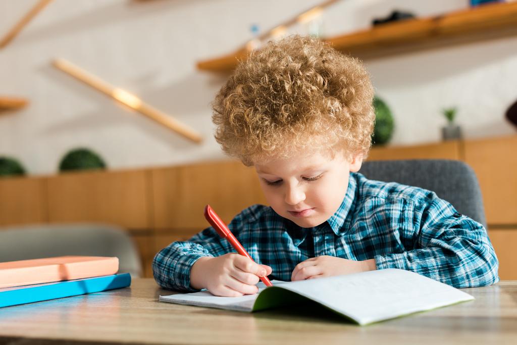 niño inteligente escribiendo en cuaderno cerca de libros en la mesa
  - Foto, imagen