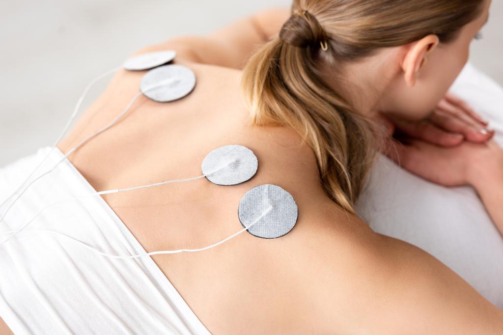 Vista de ángulo alto de la mujer joven con electrodos en la espalda durante el tratamiento de electrodos sobre fondo gris
 - Foto, Imagen