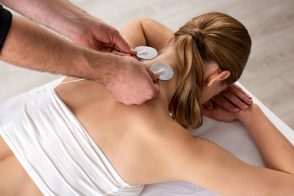 Visão de alto ângulo do terapeuta segurando eletrodos de eletroestimulação perto do pescoço da mulher no sofá de massagem
 - Foto, Imagem