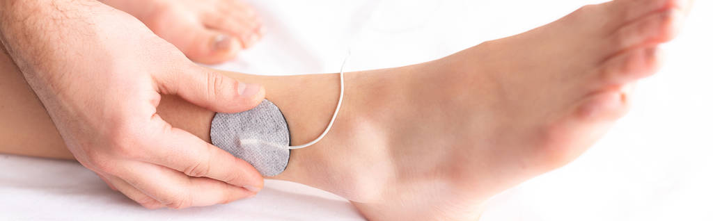 Gewassen weergave van therapeut instelling elektrode op been van de patiënt op massage bank, panoramisch schot - Foto, afbeelding
