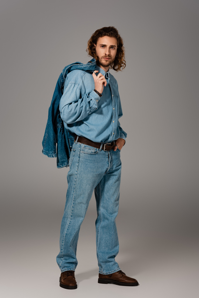 knappe en stijlvolle man in denim shirt en jeans holding jas op grijze achtergrond  - Foto, afbeelding