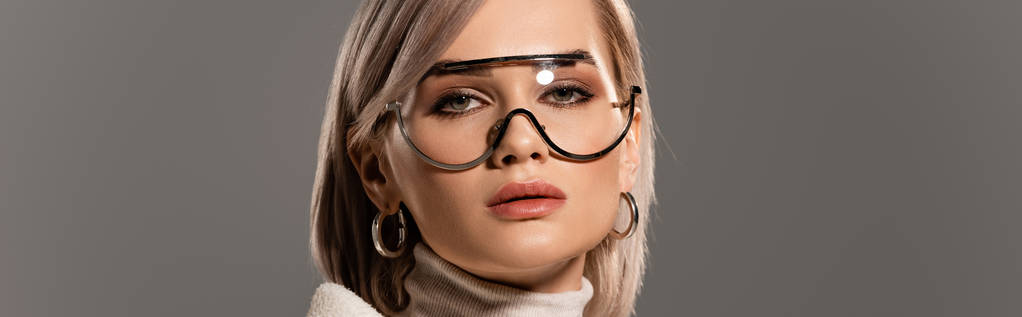 plano panorámico de mujer atractiva en gafas y pendientes mirando cámara aislada en gris
 - Foto, Imagen
