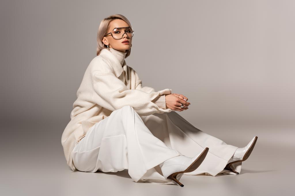 ελκυστική γυναίκα σε λευκό παλτό και παντελόνι κάθεται σε γκρι φόντο  - Φωτογραφία, εικόνα