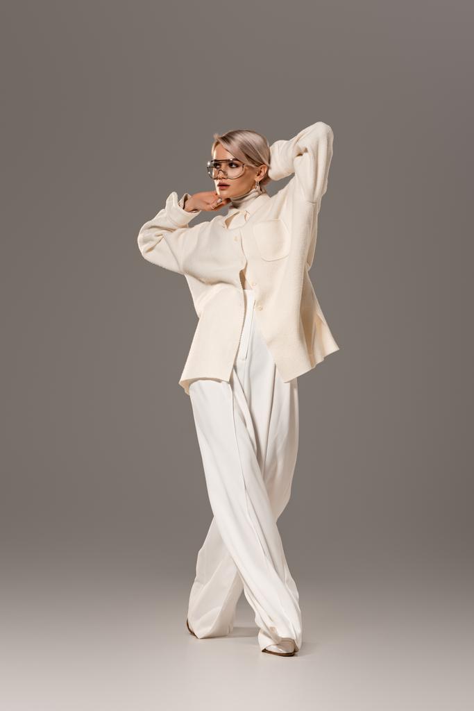 ελκυστική γυναίκα σε λευκό παλτό και παντελόνια κοιτάζοντας μακριά σε γκρι φόντο  - Φωτογραφία, εικόνα