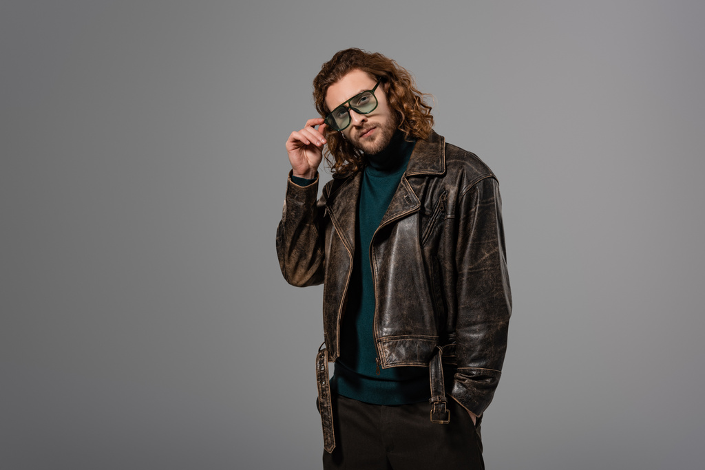 handsome man in leather jacket touching glasses and looking at camera isolated on grey  - Photo, Image