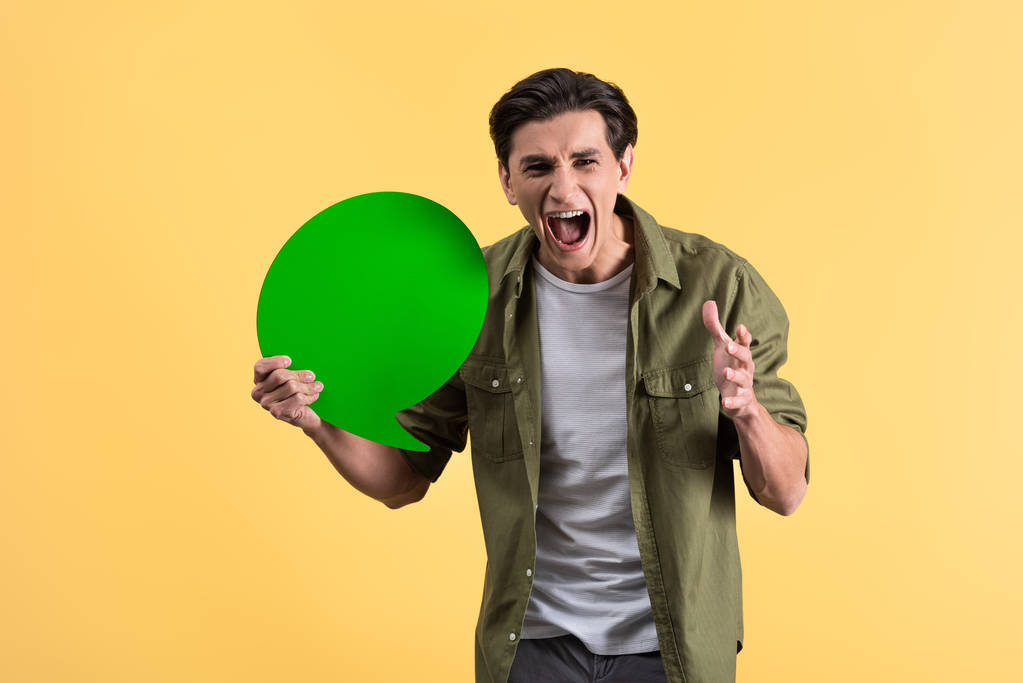 aggressive man shouting and holding green speech bubble, isolated on yellow - 写真・画像