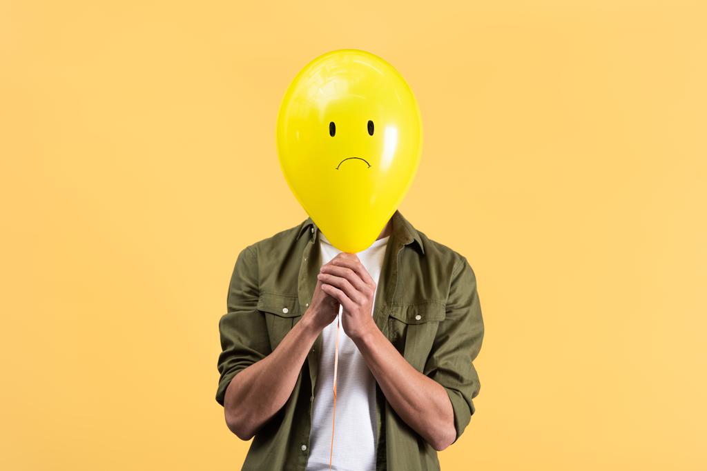 νεαρός άνδρας κρατώντας λυπημένο μπαλόνι μπροστά από το πρόσωπο, απομονωμένος στο κίτρινο - Φωτογραφία, εικόνα