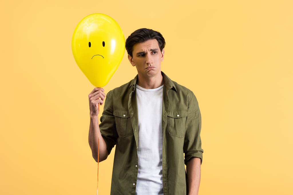 расстроенный молодой человек держит грустный воздушный шар, изолированный на желтый
 - Фото, изображение