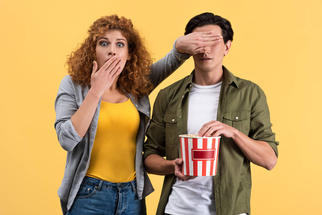 chica asustada viendo la película mientras cierra los ojos a novio con cubo de palomitas de maíz, aislado en amarillo
 - Foto, imagen