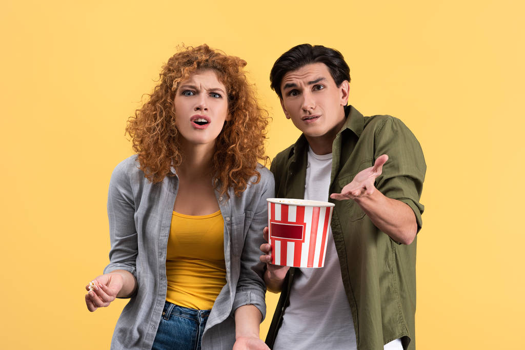 pareja irritada viendo mala película con cubo de palomitas de maíz, aislado en amarillo
 - Foto, imagen