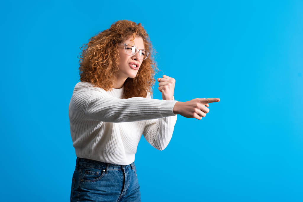 aggressive redhead girl in eyeglasses pointing and holding fist, isolated on blue - Fotoğraf, Görsel