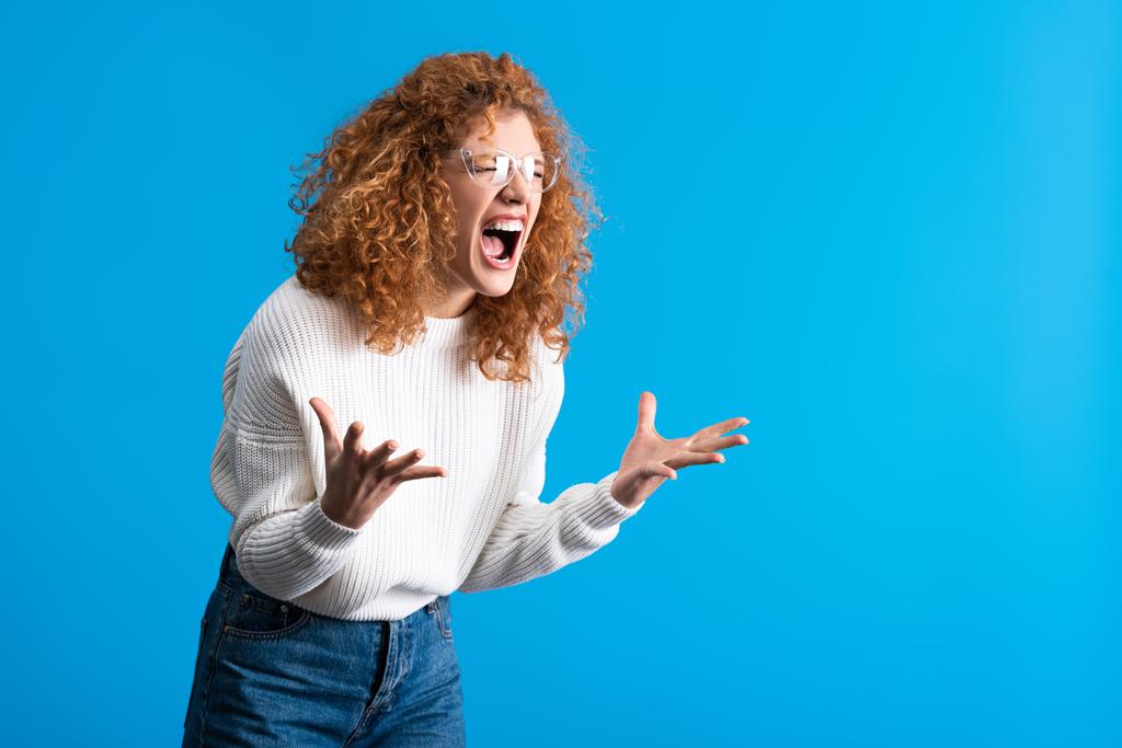 angry shouting redhead girl in eyeglasses, isolated on blue - Photo, Image