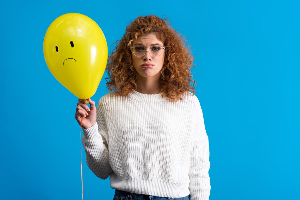triest meisje houden gele ballon met boos gezicht, geïsoleerd op blauw - Foto, afbeelding