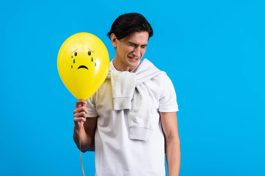 triste joven sosteniendo amarillo llorando globo, aislado en azul
 - Foto, Imagen