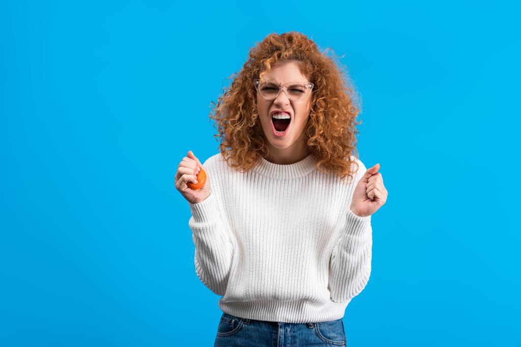 aggressive yelling redhead girl in eyeglasses squeezing stress ball, isolated on blue - Fotoğraf, Görsel
