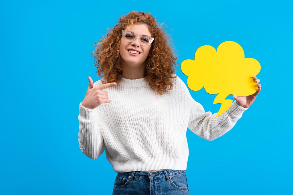 happy woman pointing at empty speech bubble in shape of cloud, isolated on blue  - 写真・画像