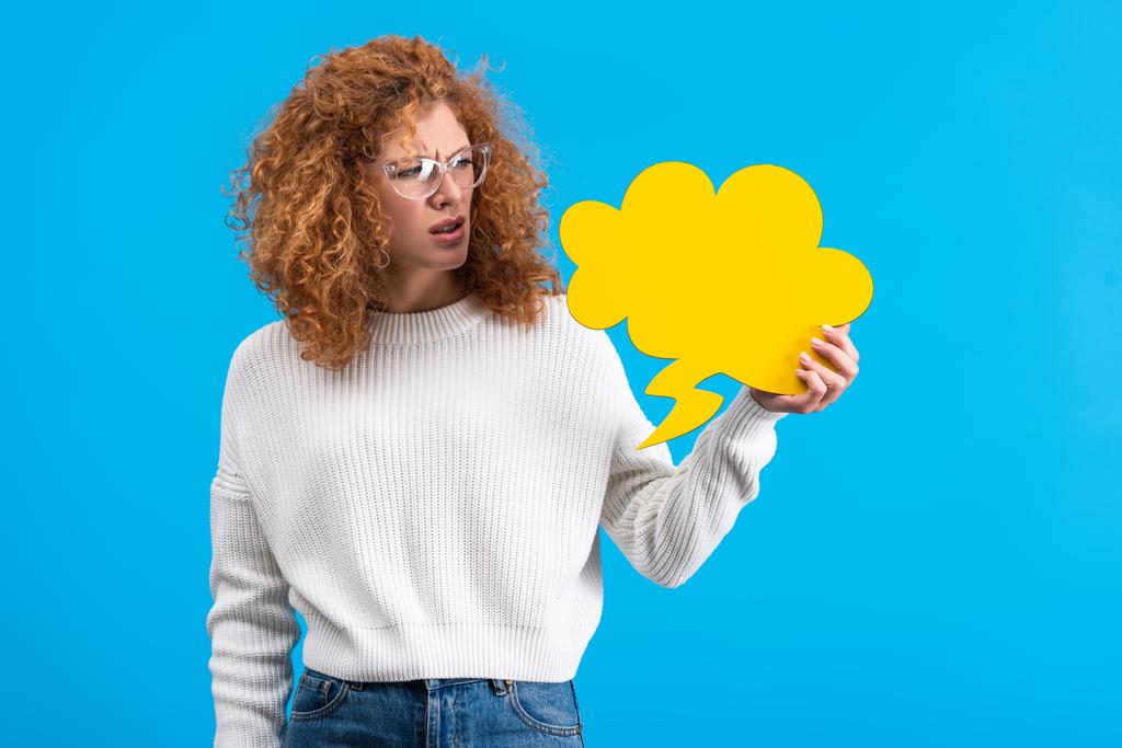 skeptical woman in eyeglasses looking at empty speech bubble in shape of cloud, isolated on blue  - 写真・画像