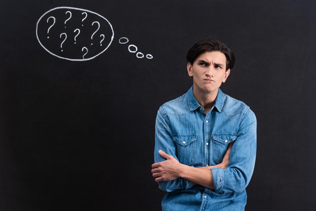 pensive young man with question marks in thought bubble on blackboard - 写真・画像